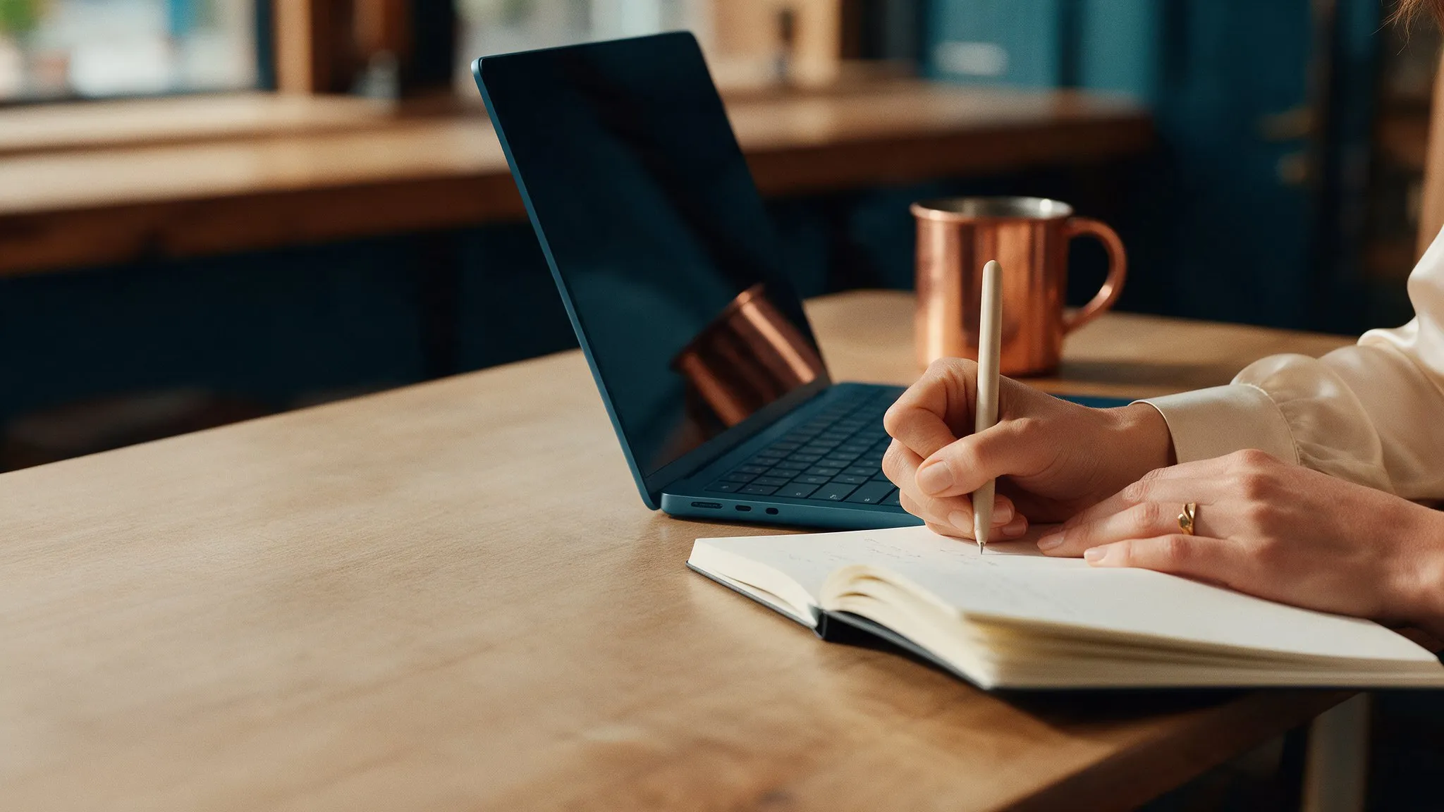 Mãos escrevendo em um caderno ao lado de um notebook e de uma caneca de cobre